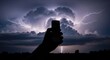© Video3 - Silhouette of a hand holding a smartphone against a dramatic stormy sky with lightning.