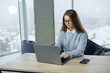 © DSMT - Woman in sweater works on laptop at table in modern office with city view on a snowy day during winter season