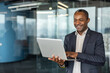 © Liubomir - Smiling businessman holding a laptop, typing and looking down at the screen, reflecting happiness and engagement while working remotely in a contemporary glass office environment