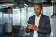 © Liubomir - African american businessman standing in a modern office, concentrating while using a mobile phone, representing professional communication, technology, and connectivity