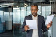 © Liubomir - Serious african american businessman standing in a modern office, concentrating while reviewing multiple business documents and comparing information for critical decision-making