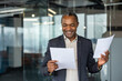© Liubomir - Experienced african american businessman smiling confidently while reviewing documents and agreements in a modern office, symbolizing success, achievement and professional satisfaction