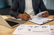 © Liubomir - Person with dark skin tones signing a formal legal agreement on a desktop with a laptop and various white paper charts, symbolizing important corporate decisions and administrative tasks