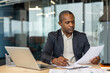 © Liubomir - Mature afro executive concentrating on legal documents at his desk, reviewing contracts and making notes beside a laptop in a bright modern office, focused and professional