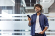 © Tetiana - Portrait of a young Indian smiling man standing indoors, holding a phone with his hand in his pocket and looking at the camera