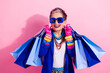 © deagreez - Vibrant retired woman shopping in bold blue jacket with colorful accessories smiles while carrying multiple blue shopping bags against a pink background
