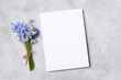 © nikavera - Blank invitation or greeting card mockup placed on a textured gray surface beside a small bouquet of blue flowers tied with twine for design purposes