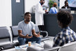 © DC Studio - Two male clients sit together in hospital waiting room, speaking before scheduled medical checkups. African American patient preparing for doctor appointment while waiting in clinical lobby.