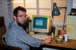 © pressmaster - Portrait of man with beard and glasses sitting at desk using vintage desktop computer, turning head toward camera, hands on keyboard, office setting visible