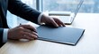 © Aqeela - Close up of a businessman in a dark suit holding a black folder and a pen while working near a modern laptop computer.