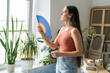 © DimaBerlin - Woman suffering from overheating at home without air conditioner, stands beside window waving hand paper fan for cooling herself. Female in need of fresh air, overwhelmed by summer heat in apartment.