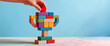 © athitat - Close up of a hand placing the final red block on a wooden toy trophy. Concept of building success, creative leadership, and step-by-step progress on a blue background with copy space.