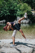 © qunica.com - Two friends perform a partner lift on a rocky river shore during a sunny day. An active fitness moment outdoors with nature in the background.