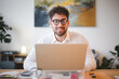 © Studio Marmellata - A man with curly hair and glasses smiles while working on his laptop at a desk.