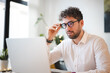 © Studio Marmellata - A man with curly hair adjusts his glasses while working on his laptop. He is wearing a white collared shirt and bracelets on his wrist.