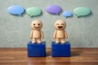 © Jumpei - 木製のブロックに乗って笑顔で会話を楽しむ木の人形とカラフルな吹き出しのコンセプト