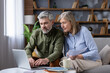 © Liubomir - Mature couple on sofa in cozy living room using laptop and papers to review bills, budget and retirement plans, focused on savings, mortgage and investments for future