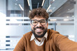 © Liubomir - Young professional man with beard and curly hair wearing glasses, smiling brightly while making a video call or taking a selfie in a contemporary office environment