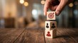 © Muhammad - A human hand carefully placing a wooden cube with a red target icon onto a stack of two wooden cubes featuring black and red people icons on a warm rustic wooden desk in