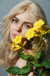 © VICHIZH - Portrait of a young woman with long blonde hair holding yellow flowers against a soft gray background, evoking a sense of freshness and beauty.