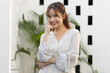 © 220 Selfmade studio - Portrait of cheerful beautiful asian woman wearing white stylish dress smiling at camera with hand on chin standing near green plant and white wall background looking happy confident and positive