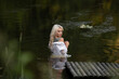 © shymar27 - Female model with long blonde hair wearing a sheer white shirt poses in a tranquil pond surrounded by greenery and wooden structures on the water's edge