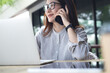© tippapatt - Asian woman talking on mobile phone while working on laptop computer on tablet at outdoor cafe, surfing internet, remote work, online business casual using smartphone