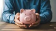 © thenort - Person in blue shirt cradles a pink ceramic piggy bank with both hands over wooden table