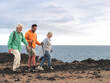 © luciano - Three people from different generations enjoy a coastal hike along a rocky shoreline, walking together with trekking poles under a cloudy sky by the sea.