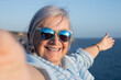 © luciano - Close-up portrait of a smiling senior woman wearing sunglasses taking a selfie by the sea at sunset. Happy and carefree, she enjoys a summer vacation with ocean in the background
