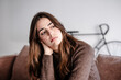 © luciano - Portrait of serious and pensive young woman with long brown hair sitting on couch at home looking away