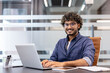 © Tetiana - Portrait of a young Indian man in glasses and a shirt sitting at a desk in an office, working on a laptop and looking at the camera