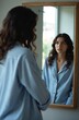 © Pete - Woman in blue striped pajamas looks in mirror reflection. Natural morning light fills room. She appears relaxed and thoughtful, embracing personal style and self care.