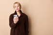 © SHOTPRIME STUDIO - Mature woman in brown coat looks at smartphone with neutral expression. Isolated on beige background. Concept of technology use, communication, and casual lifestyle for older adults.