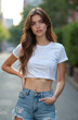 © Viktor - Young woman wearing casual summer street style outfit. She poses confidently outdoors with hands on hips and in pockets, showing off denim shorts and white crop top.