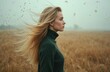 © Maryna - Blonde woman with hair blowing in wind stands in golden field wearing dark green sweater. Outdoor natural scene with soft light and distant trees.