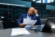 © Anastasiya - Mature black businessman reviewing printed document while touching forehead in modern office. Serious executive analyzing paperwork and making strategic decisions