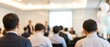 © CuteBee - A blurry view of business professionals attending a corporate event or seminar, with a presenter at the front and decorative balloons on the side.