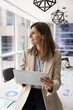 © fizkes - Confident businesswoman in modern meeting room holds tablet while looking into distance with thoughtful smile, ponder on corporate strategy or creative start-up ideas. Break, productivity, leadership
