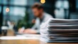 © dmmarychev - Stack of white paper documents on a wooden desk with a blurred office worker in background