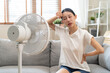 © Pormezz - Suffer summer heat wave stroke, hot weather, tired asian young woman sitting on couch in living room without air-conditioner, using electric fan, wave fan to cool for ventilation when high temperature