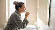© mangolovemom - Woman toothbrushing bathroom mirror hygiene morning routine in bathrobe near sink, smiling while cleaning teeth with toothbrush, natural light, home wellness