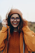 © SHOTPRIME STUDIO - Smiling woman lifestyle wearing glasses, knit hat, and orange jacket outdoors in cool weather. Happy young female enjoying nature, casual fashion, and positive mood on autumn day.