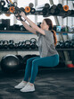 © MZaitsev - Young woman exercising in gym using suspension straps for squat workout, focusing on strength and balance while training among kettlebells and fitness equipment indoors