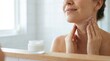 © Stitch - Woman applying anti-aging firming cream to her neck and jawline for skin care in front of a mirror.