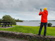 © mark_gusev - A man in a red jacket is taking a picture of the lake. The sky is cloudy and the water is calm. Travel and tourism. Calm and relaxing mood.
