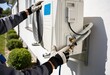 © Netsai - A technician wearing gloves is installing or servicing an outdoor climate control unit on the exterior wall of a residential building, connecting pipes.