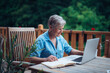 © Halfpoint - Mature woman sitting on balcony with laptop, working remotely.
