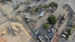 © DawDunia - Aerial view of a sprawling industrial storage yard and construction site with excavators, tractors, stacked pipes and concrete blocks amid dirt piles and open-air building material storage