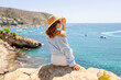 © maxbelchenko - A young woman sits on a cliff, wearing a hat, overlooking the seascape. A rear view of a woman feeling free, enjoying a sunny day by the sea. Concepts of nature, freedom, and weekends.